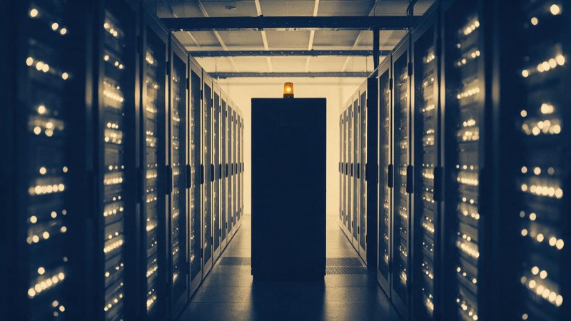 A single dark cabinet among rows of lit ones in a data centre aisle, a lone amber light above it
