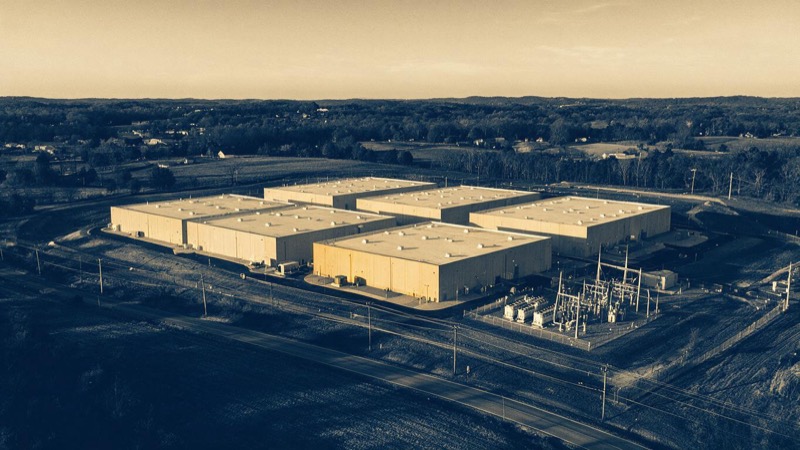 Aerial of a hyperscale data centre complex in northern Virginia at golden hour, windowless sand-coloured buildings, a substation and cooling units beside a quiet rural road