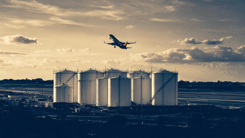 A fuel storage depot of tall white tanks beside an airport at sunset, a jet climbing out above them