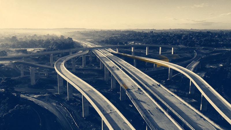Aerial view of a motorway interchange under construction outside Sydney, concrete pillars and curved ramps half-finished, in early morning haze