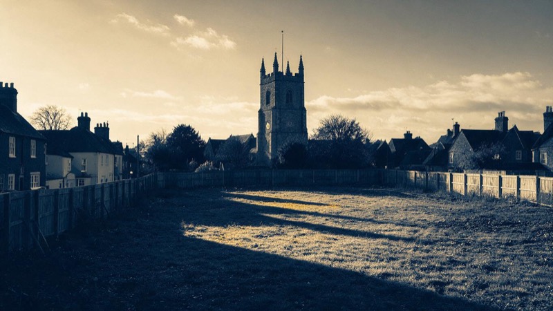 An empty development plot in an English market town with a church tower behind it, long shadows falling across the grass at golden hour