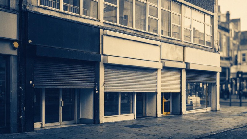 A high street of commercial shopfronts and first-floor offices in a city centre, shutters half raised, photographed in early morning