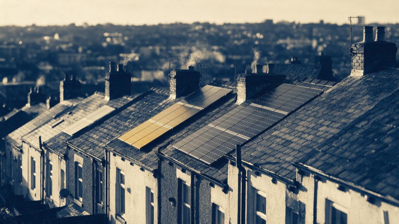 Rooftop view of terraced houses in Leeds with a few roofs carrying new solar panels, the rest bare slate, chimneys smoking faintly in cold clear morning light