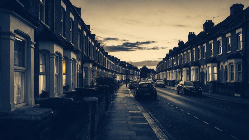 A long street of terraced brick houses in a London suburb at dusk, windows lit and parked cars along the kerb, photographed from the far end of the pavement