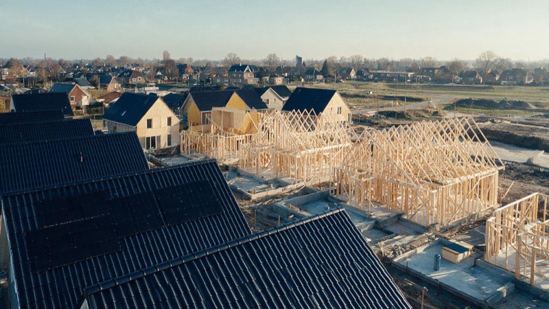 Aerial view of a new housing estate half built on the edge of a Dutch town, finished roofs beside bare timber frames and concrete foundations, in flat morning light