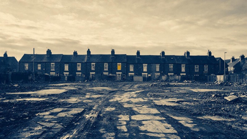 A cleared brownfield site on the edge of Manchester, mud tracks and broken concrete slabs with a row of old brick terraces behind, under grey morning cloud