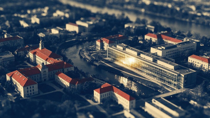 Late-afternoon aerial of a university science campus, red-tiled roofs, a river bend and a long glass laboratory building catching the sun