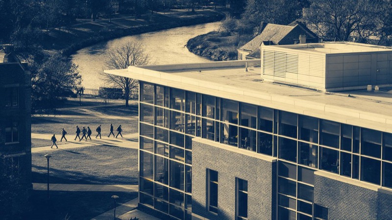A university research building of glass and brick at golden hour, students crossing the quadrangle in the distance and a river bend behind the campus