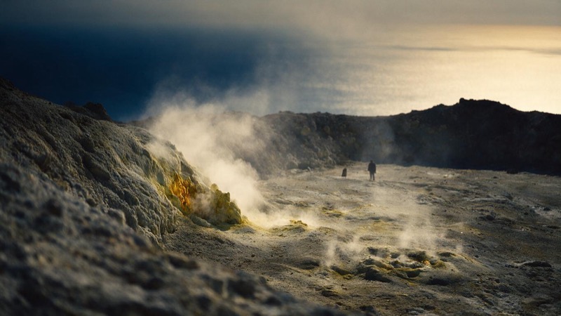 Steam rising from yellow-stained vents on a barren crater floor in southern Italy, a lone figure far off and the hazy sea beyond the rim