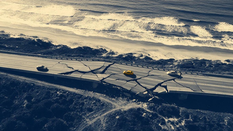 Aerial view of a long coastal highway buckled and split by an earthquake, the Pacific beyond, a few stopped vehicles small on the cracked road