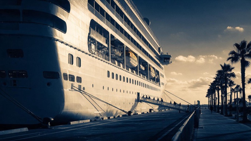 A large white cruise ship at its berth beside a palm-lined promenade at dusk, passengers small on the gangway