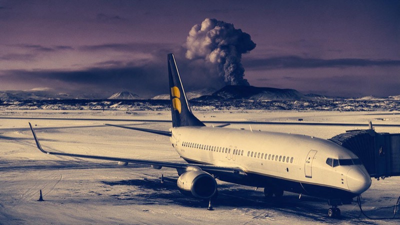 An airliner parked on a snowy apron at an Icelandic airport at dusk, a plume of ash rising from a distant volcano under a violet sky