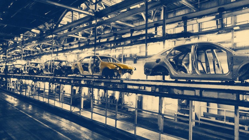 Wide view of a car assembly line in Puebla with painted bodies hanging from an overhead conveyor and a conspicuous empty gap in the line, bright factory light
