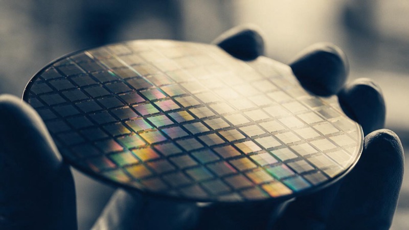 Extreme macro of the iridescent grid of dies across a silicon wafer, rainbow reflections sliding across the surface, held at an angle in a gloved hand against soft grey