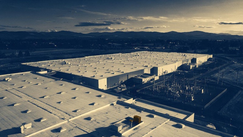 Aerial of a large data centre complex in Oregon at dusk, vast roofs and a substation, mountains in the distance