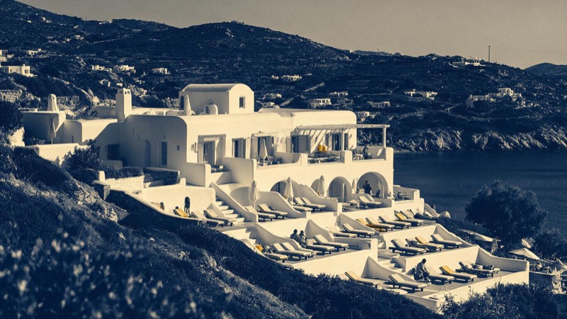 A white resort hotel stepping down a hillside beside a turquoise bay on a Greek island, sunbeds in rows on the terraces, in bright mid-morning sun