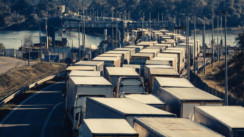 Aerial view of a long queue of freight lorries inching toward a river border bridge in mid-morning, white cabs lined up for a mile
