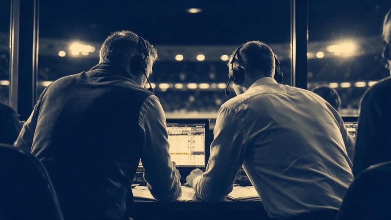 A bookmaker's trading room at night in Gibraltar, long desks with angled monitors glowing as soft blurred light and a stadium's floodlights visible through the window