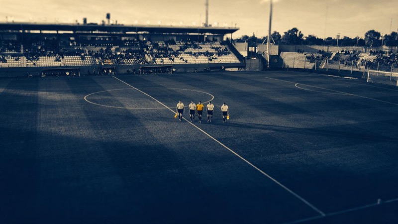 Referees and linesmen walking a pitch before kick-off at a regional stadium in Argentina, seen from far above in late light, stands half full