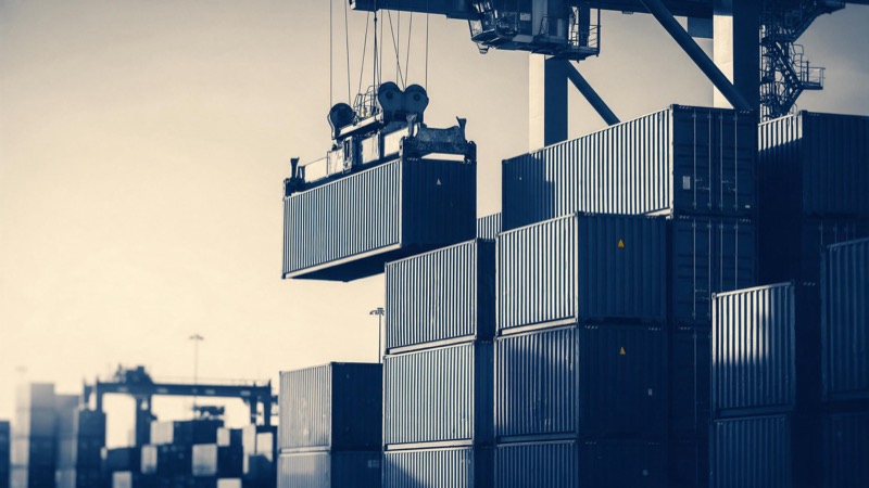 stacked shipping containers at a port, a gantry crane lifting one