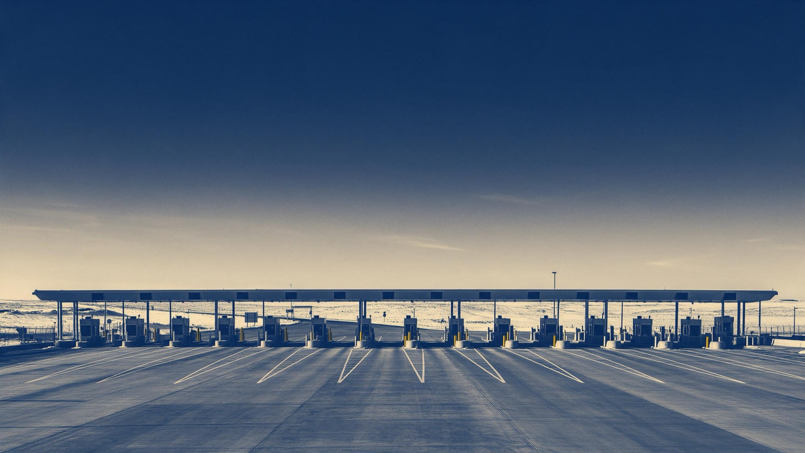 A wide border crossing plaza with rows of inspection booths under a long canopy, empty lanes and snow-dusted prairie behind
