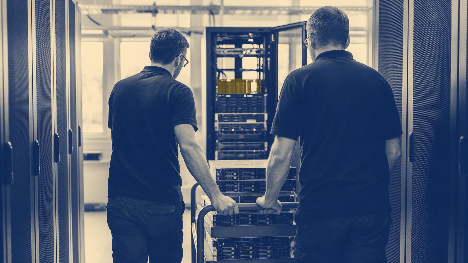 Two data centre engineers seen from behind wheeling a cart of new servers down a long aisle toward an open rack, hands on the handle