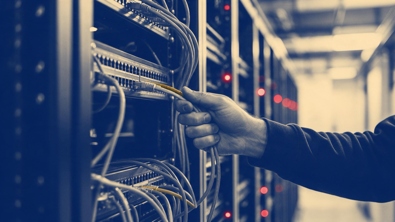 A technician's hand pulling a network cable from a rack as a row of red indicator lights glows along the aisle