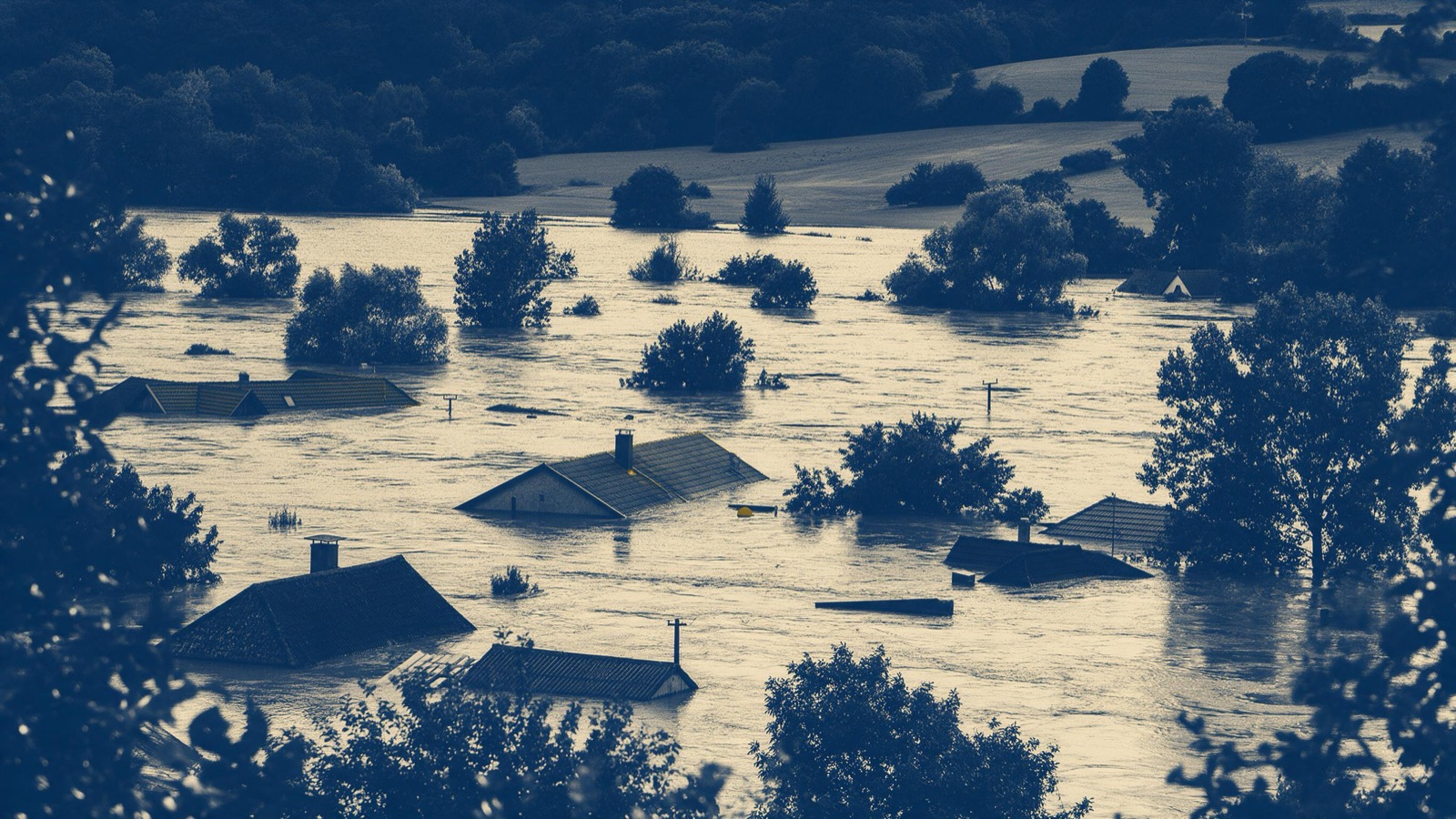 A rural valley under heavy flood water seen from a hillside after a storm, roofs and treetops poking above the brown surface