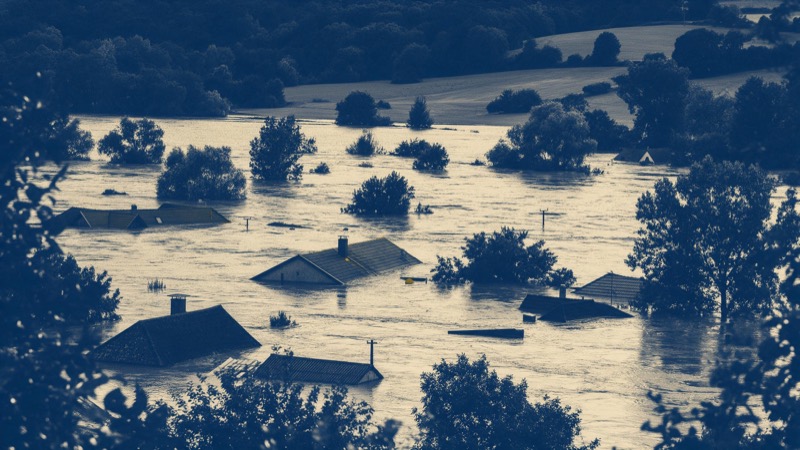 A rural valley under heavy flood water seen from a hillside after a storm, roofs and treetops poking above the brown surface