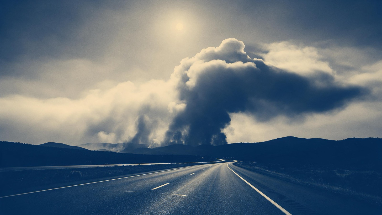 A vast wildfire smoke column rising over distant hills at midday, the sun dimmed to a pale disc above an empty highway