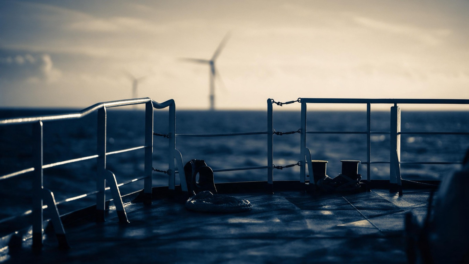 Two technicians in survival suits seen from behind on a turbine platform, hands on the rail, looking out across a field of turning rotors