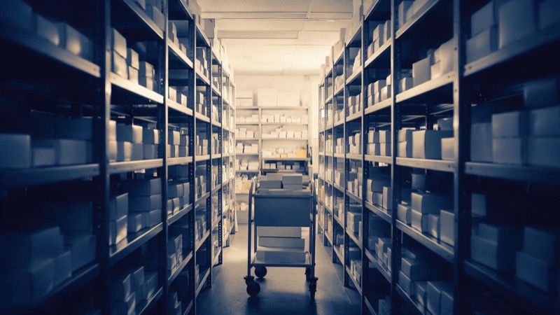 A hospital pharmacy store room lined with plain white boxes on steel shelves, lit by cold overhead light, with a trolley parked in the aisle