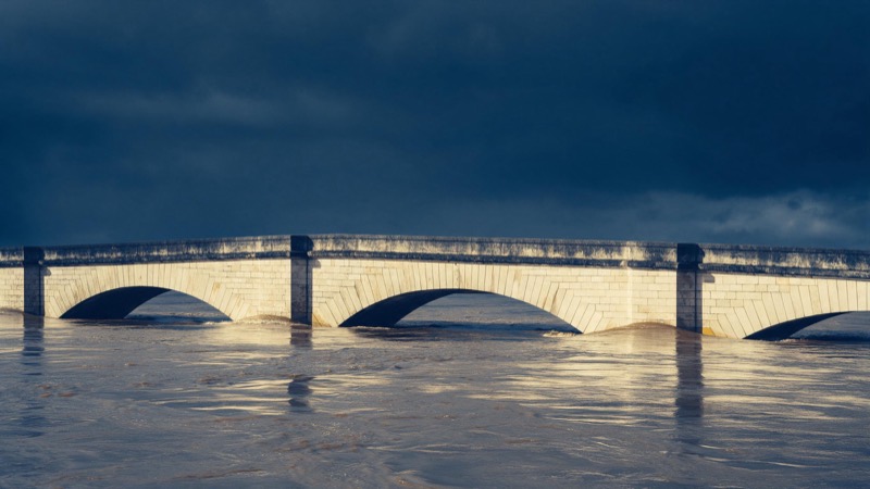 A swollen river brimming at the top of its banks, brown water pressing against a stone bridge under a heavy grey sky
