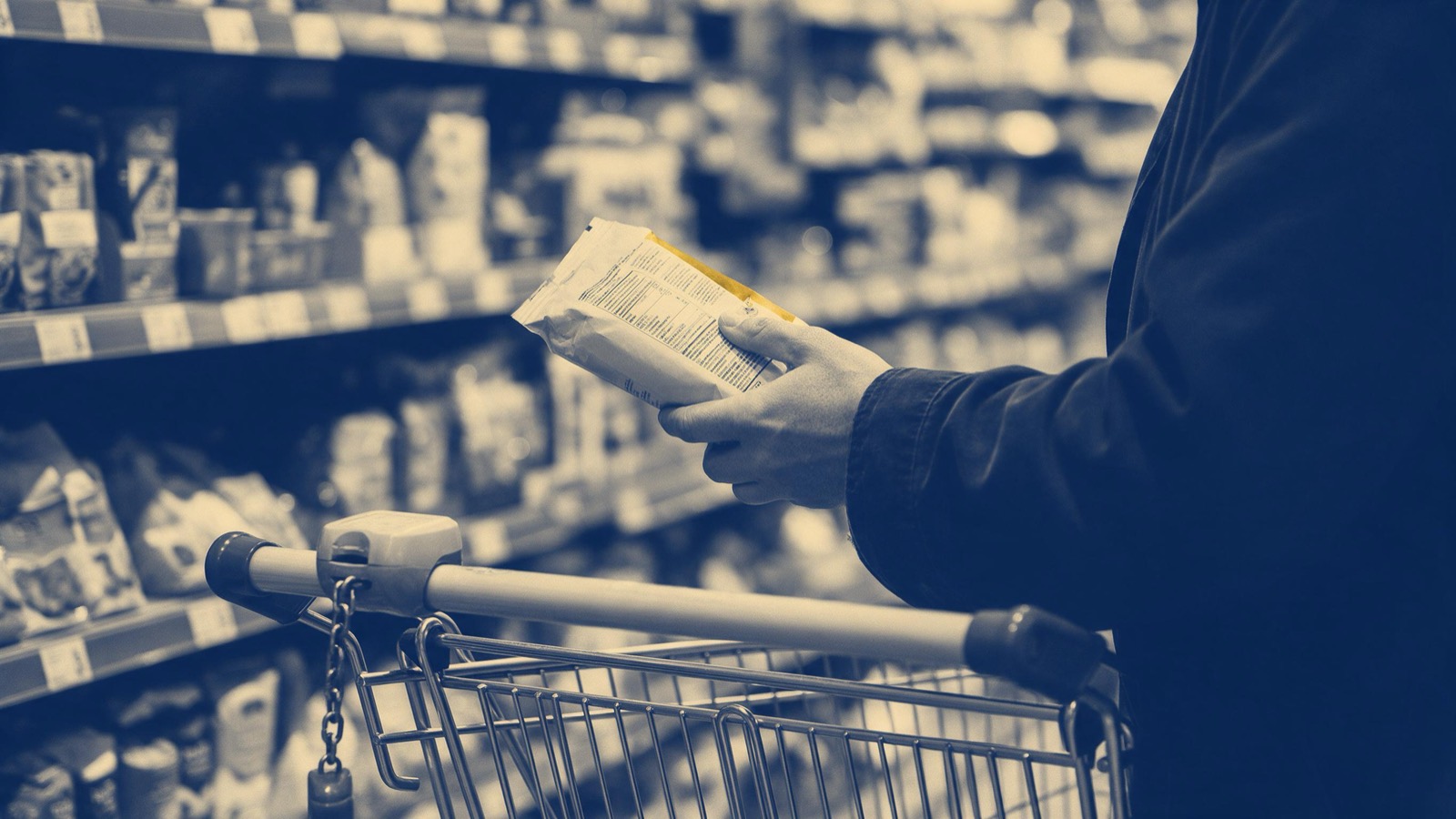 A shopper's hand turning a plain packet over in a supermarket aisle to inspect its back, trolley in the foreground, face out of frame