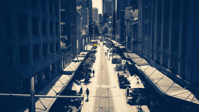 A pedestrian city-centre street between tall buildings seen from a high balcony at midday, a tram line down the middle and café awnings along the sides