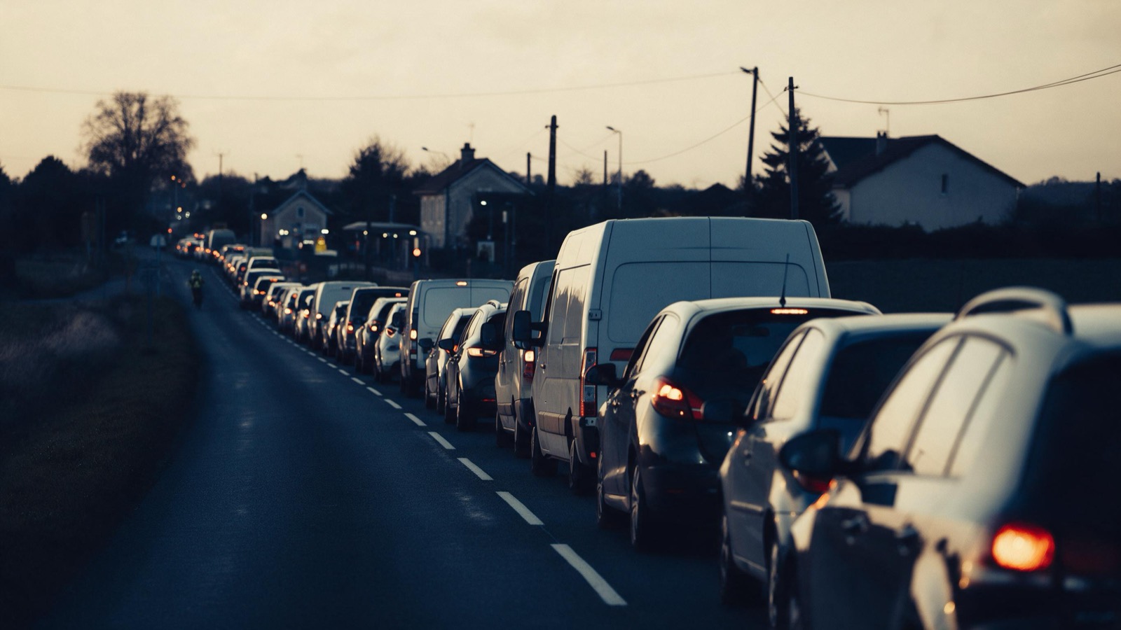 A long queue of cars and vans stretched along a French country road at dusk, tail lights glowing, waiting to reach a distant station