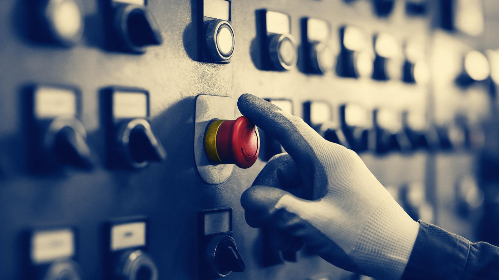 A gloved hand pressing a red emergency stop button on a factory control panel, rows of switches blurred behind it