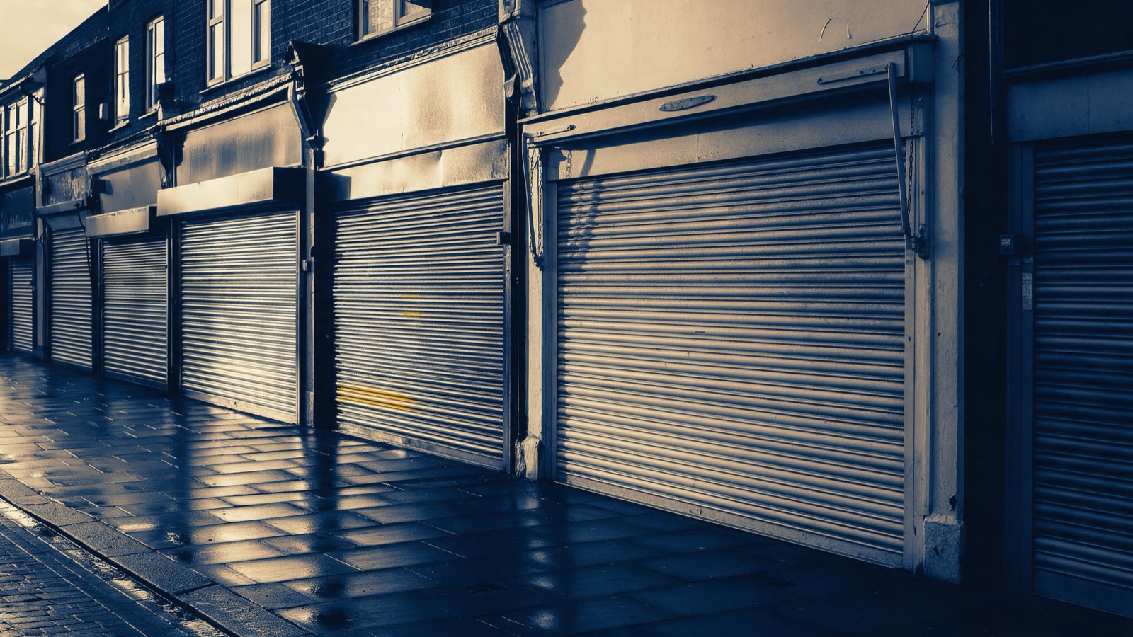 A row of shuttered shopfronts on an English high street at dusk, metal blinds pulled down and wet pavement glistening