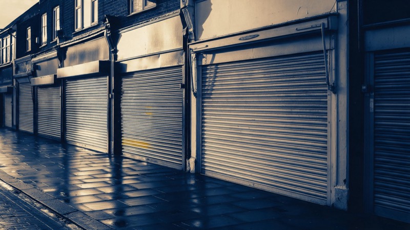 A row of shuttered shopfronts on an English high street at dusk, metal blinds pulled down and wet pavement glistening