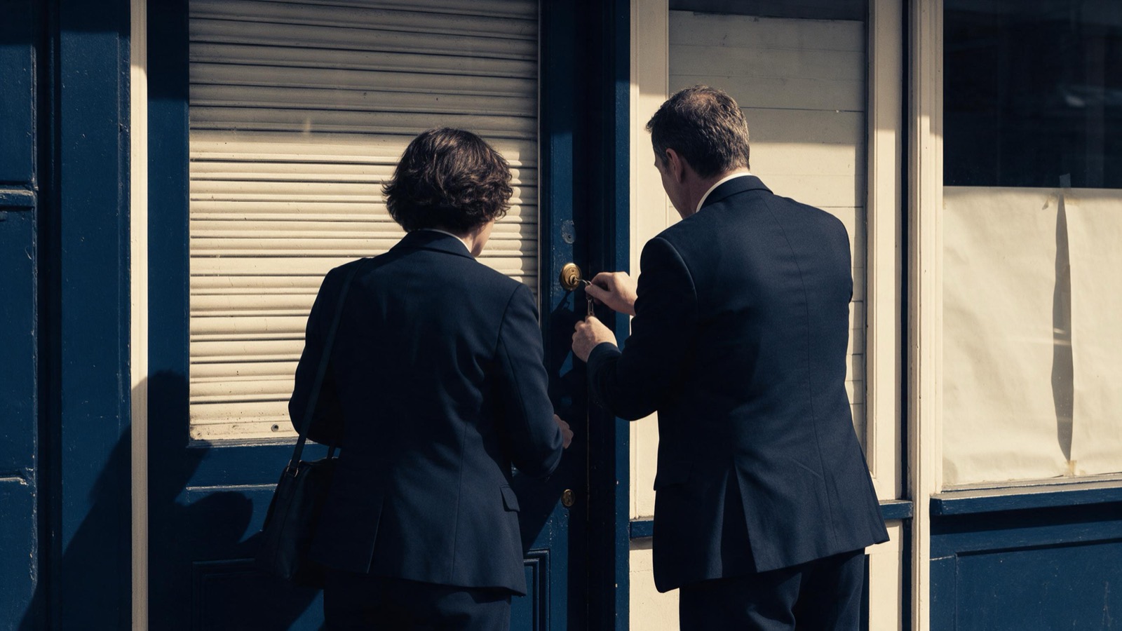 Two people in dark suits seen from behind at the door of a closed shop, one turning a key in the lock
