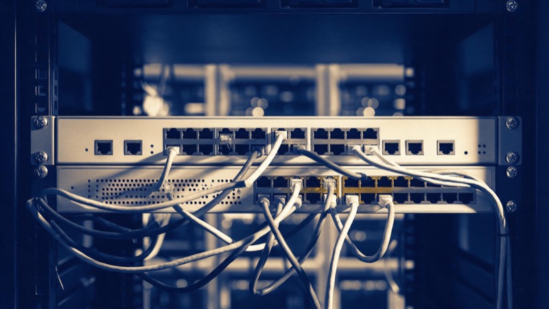 An unbranded network firewall and switch mounted in a rack, rows of ports and tangled patch cables photographed close under hard white light