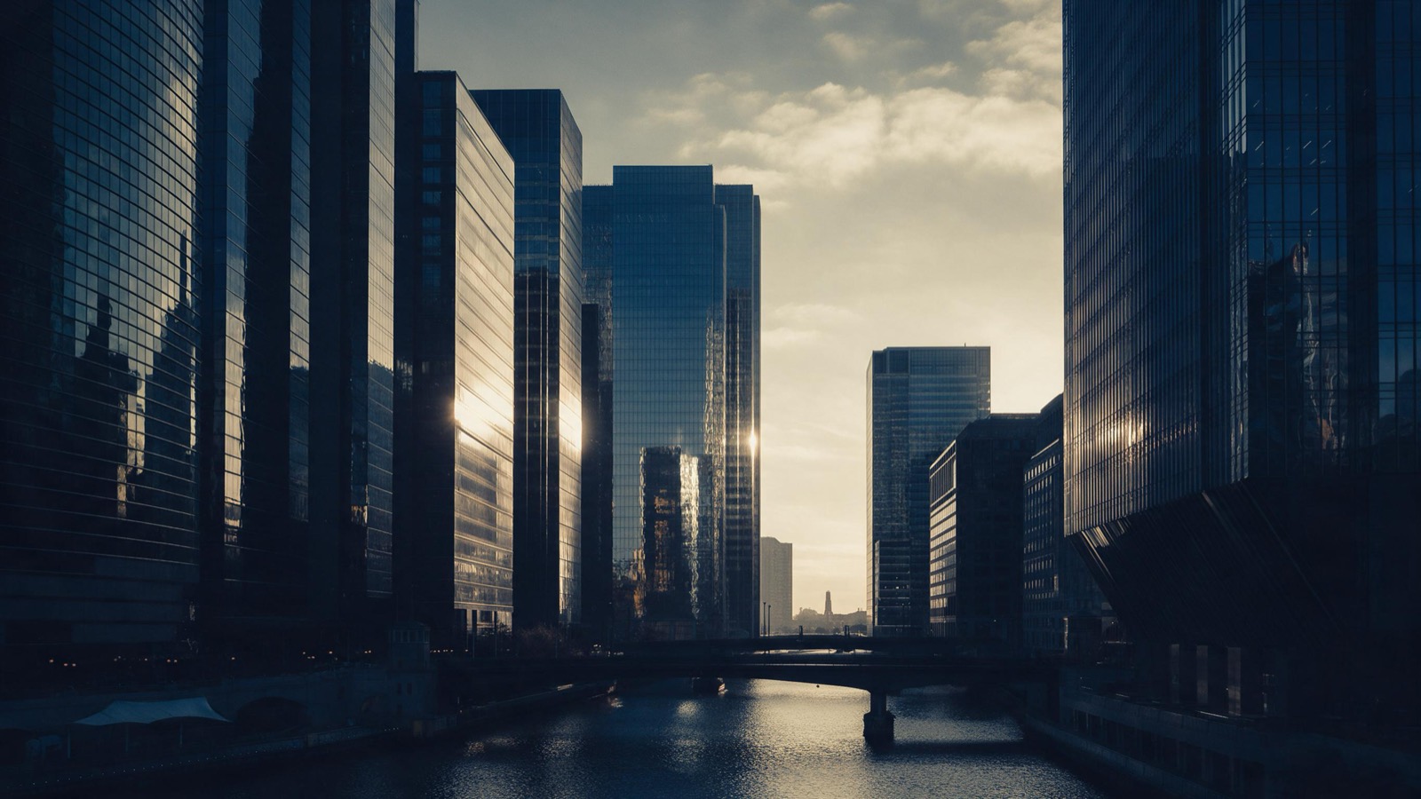 A financial district skyline at first light seen across a river, glass towers catching the sun above empty streets and quiet bridges