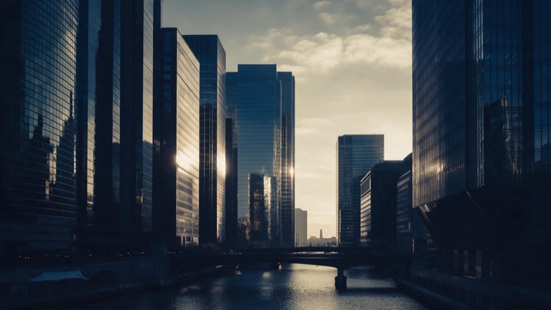 A financial district skyline at first light seen across a river, glass towers catching the sun above empty streets and quiet bridges