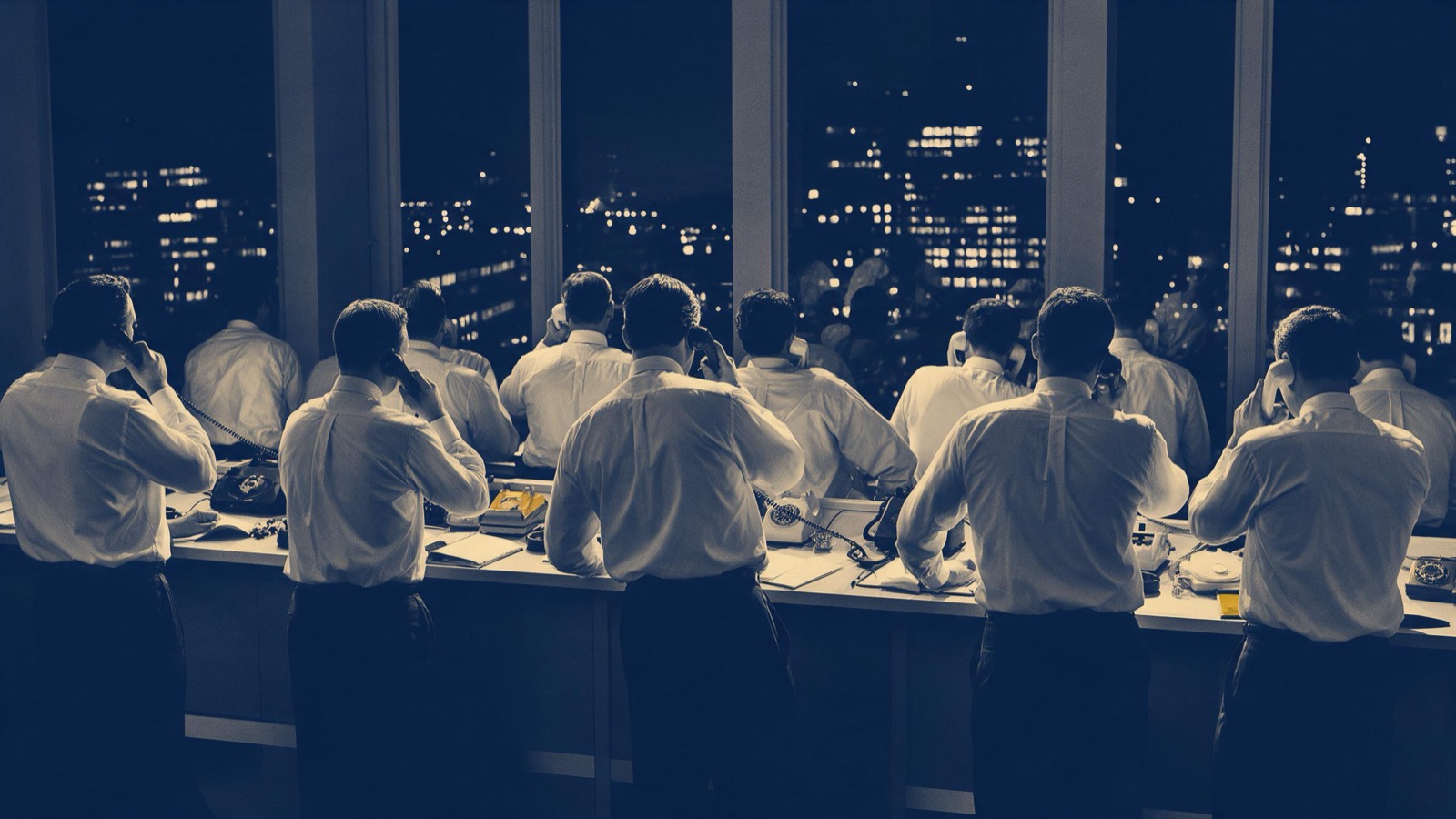 Dealers in shirtsleeves seen from behind at a long open-plan desk at night, telephones at their ears, city lights spread beyond the tall windows