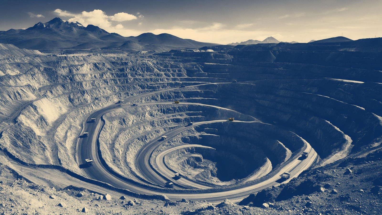 An open-pit copper mine in the high Andes seen from the rim in late afternoon, terraced rust-red walls spiralling down, tiny haul trucks crawling on the ramp