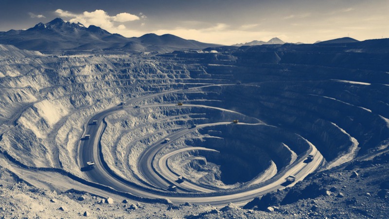 An open-pit copper mine in the high Andes seen from the rim in late afternoon, terraced rust-red walls spiralling down, tiny haul trucks crawling on the ramp