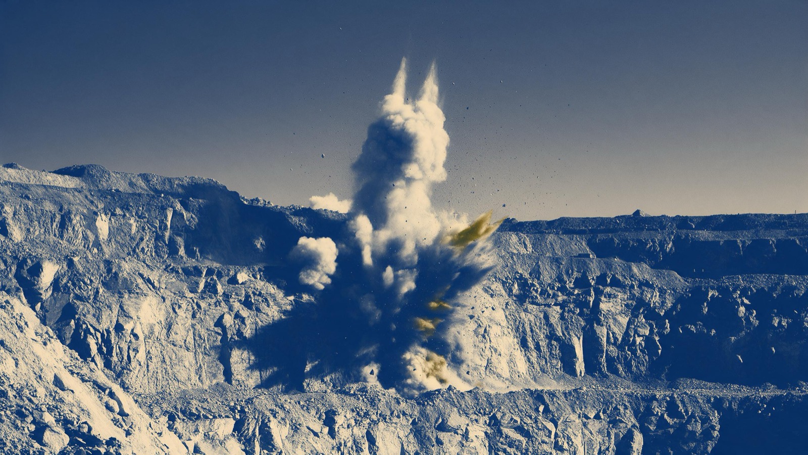 A controlled blast on the wall of an open pit seen from a safe distance, a plume of dust and rock rising against a clear sky