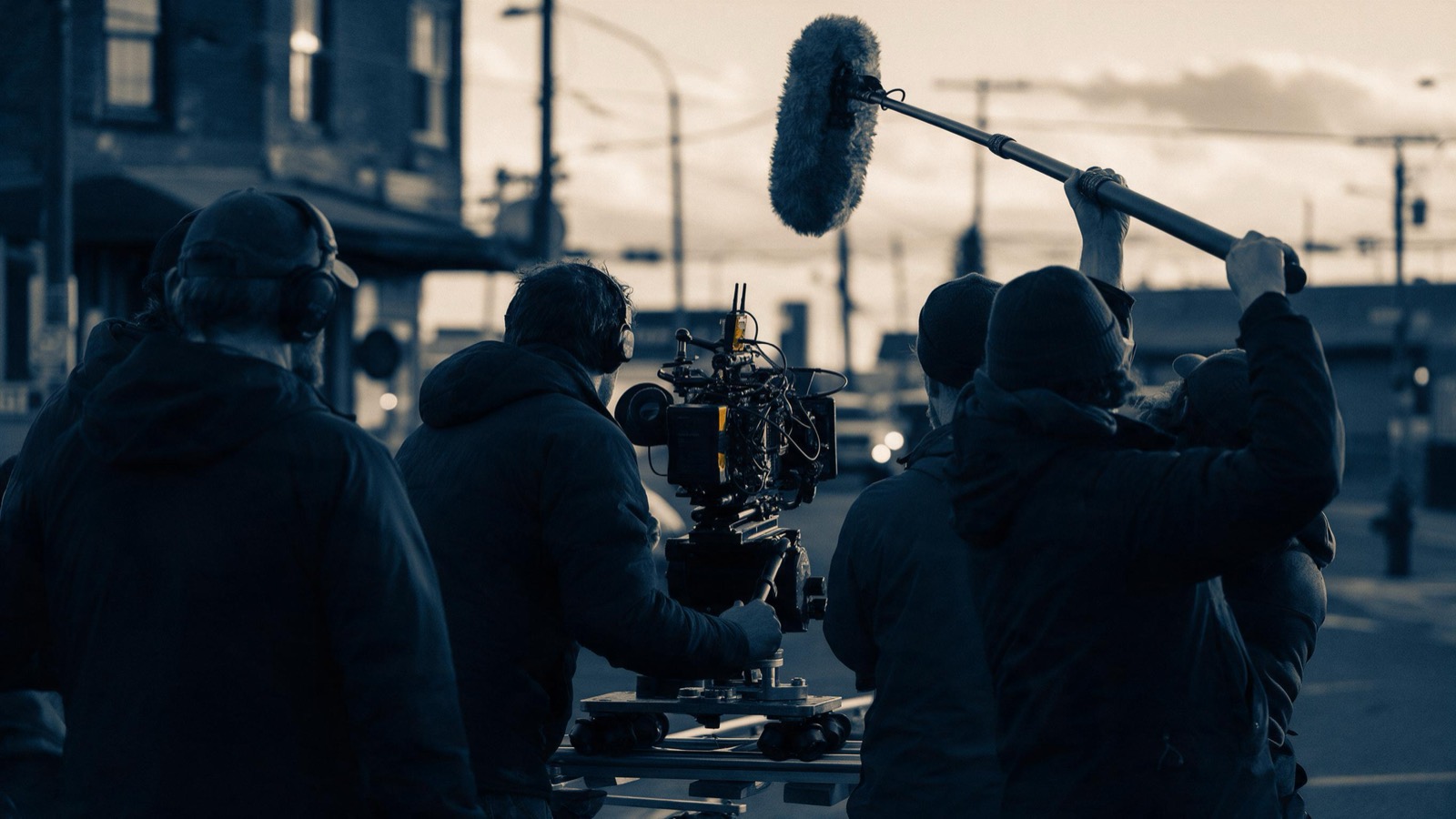 A film crew seen from behind around a camera on a dolly track in a street at dusk, a sound pole held overhead