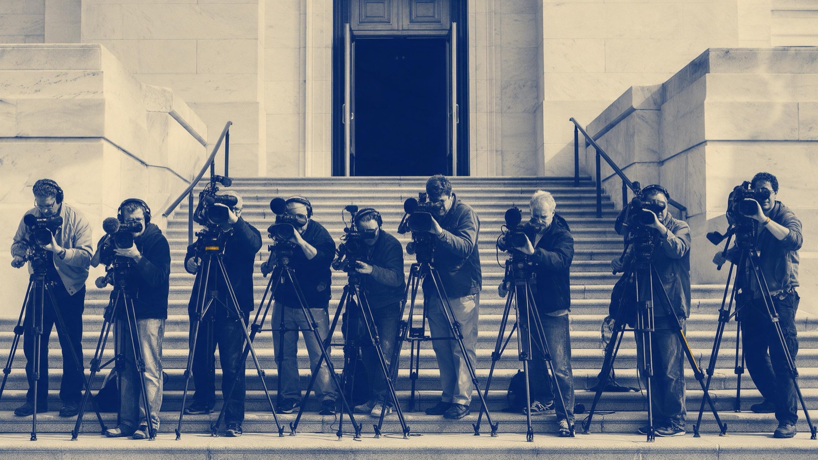 Camera operators with tripods lined up on the steps of a government building as a tall door opens at the top