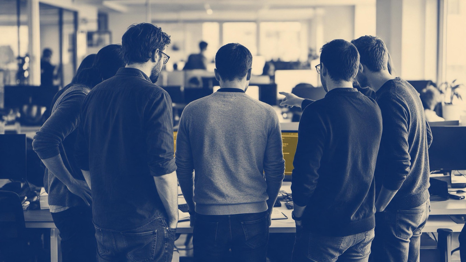 Developers seen from behind standing together around one desk in an open-plan office, one pointing at something just out of frame